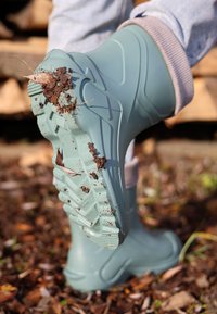 Botte imperméable vert bleu clair avec une semelle texturée, présentant de la boue et des feuilles, conçue pour une utilisation en extérieur. La tige est lisse avec une coupe ajustée.