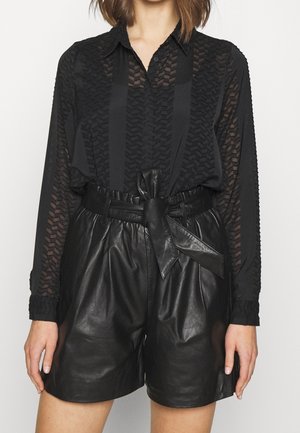 Nainen pukeutuneena mustaan läpikuultavaan kuvioituun pitkän hihan paitaan, joka on sullottu korkeavyötäröisiin mustiin nahkashortseihin, joissa on solmittu vyö.