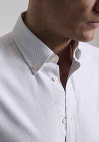 Camisa blanca de botones con finas rayas azules, fabricada en suave algodón, con un cuello clásico y sutiles detalles de botones.