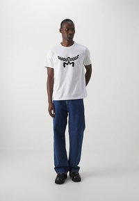 Weißes Baumwoll-T-Shirt mit schwarzem Grafikdesign auf der Vorderseite, kombiniert mit blauen Jeans und schwarzen Schuhen.