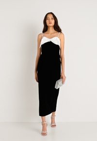 Μαύρο βελούδινο φόρεμα με λευκή σατέν λεπτομέρεια στον λαιμό, λεπτές τιράντες και εφαρμοστή σιλουέτα; συνδυασμένο με μια ανοιχτό γκρι τσάντα και τακούνια.