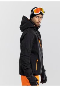 Giacca da sci nera con accenti arancioni, cappuccio, tasche con zip e polsini regolabili, abbinata a guanti neri e pantaloni arancioni.