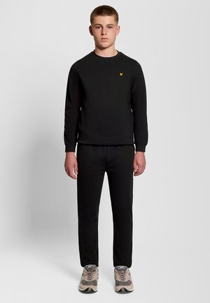 Tonåring klädd i svart sweatshirt och byxor med beige New Balance-sneakers, stående mot en enkel ljusgrå bakgrund.