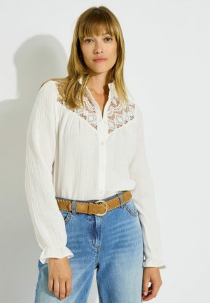 Mujer con el pelo rubio liso, vistiendo una blusa blanca de manga larga con detalles de encaje y vaqueros azules con un cinturón marrón sobre un fondo blanco.