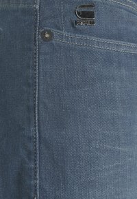 Džínové kalhoty v prostřední modré barvě s hladkou texturou, s viditelným švem kapsy, kovovým knoflíkem a logo štítkem blízko pasu.