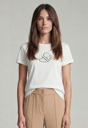 Young woman with long brown hair wearing a white t-shirt with a black abstract design and beige high-waist pants against a plain background.