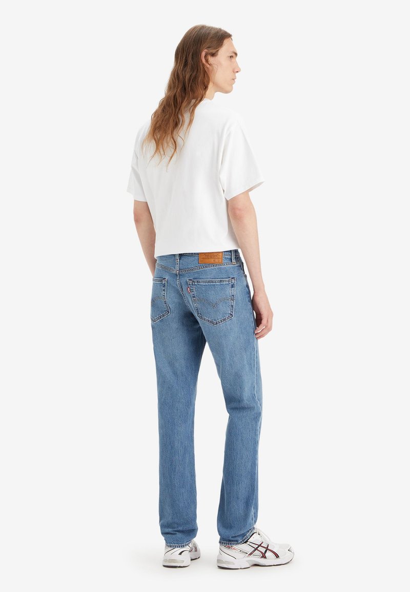 Persona con cabello largo, vistiendo una camiseta blanca, jeans azules y zapatillas blancas, de pie de espaldas sobre un fondo liso.