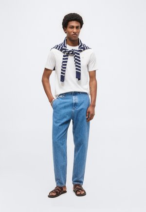 Mężczyzna o afro fryzurze, w białej koszulce, pasiastym swetrze narzuconym na ramiona, niebieskich dżinsach i brązowych sandałach, stoi z jedną ręką w kieszeni.