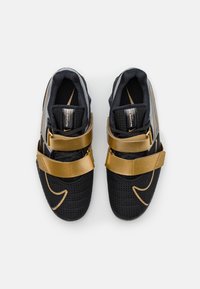 Scarpe da ginnastica nere in materiale mesh, con cinturini dorati e un design testurizzato con un logo swoosh prominente.