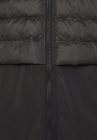 Chaqueta acolchada negra con una textura suave, que presenta una cremallera de longitud completa en el centro y un diseño a bloques de color en el pecho.