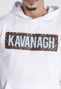 Sudadera blanca con capucha que presenta un gran parche de logo marrón con "KAVANAGH" en azul claro y motivos de diseñador repetitivos. Textura de tela suave.