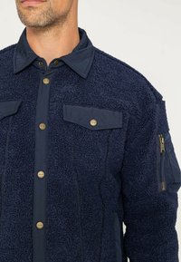 Granatowa polarowa kurtka o teksturowej powierzchni, z kontrastowym krawatem, dwoma kieszeniami na piersi oraz zamykaną na zamek kieszenią na rękawie z złotym okuciem.