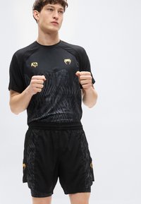 Μαύρο αθλητικό σετ που περιλαμβάνει ένα κοντομάνικο μπλουζάκι με υφή και χρυσές λεπτομέρειες, συνοδευόμενο από σορτς με πλαϊνές τσέπες που ταιριάζουν.