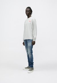 Noor mees seisab, seljas helehall pikkade varrukatega särk punase logoga, sinised teksapüksid ja hallid kõrge säärega tossud valgel taustal.