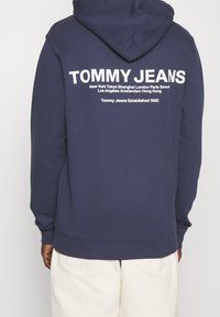 Námořnická modrá mikina s kapucí a klokanní kapsou. Na zadní straně se nachází bílý text včetně názvů měst a loga značky. Materiál z bavlněné směsi.