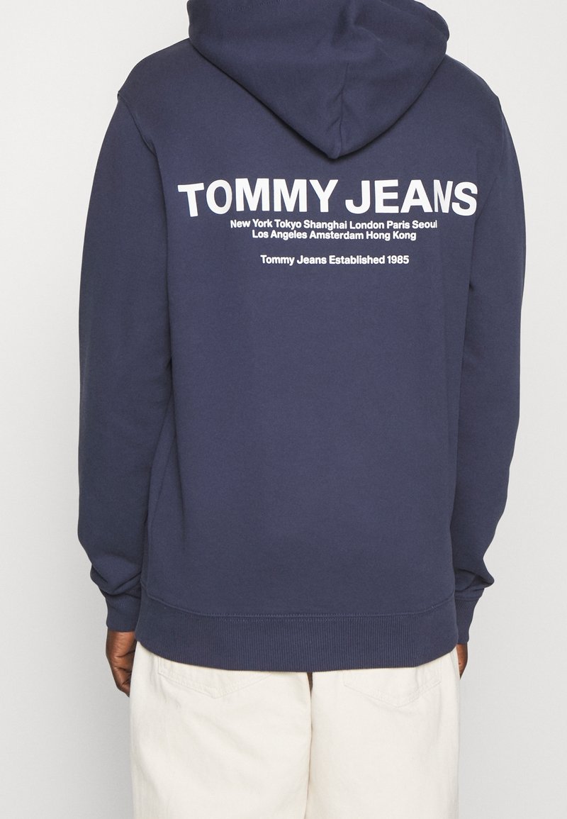 Námořnická modrá mikina s kapucí a klokanní kapsou. Na zadní straně se nachází bílý text včetně názvů měst a loga značky. Materiál z bavlněné směsi.