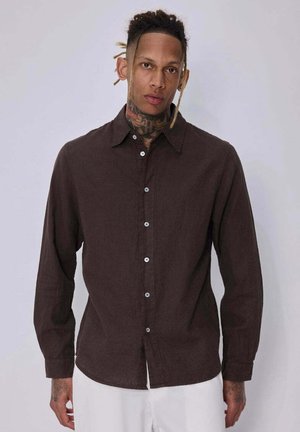 Homme avec des tatouages et des dreadlocks portant une chemise marron foncé boutonnée et un pantalon blanc sur un fond clair uni.