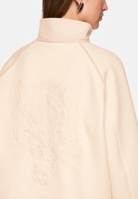 Light beige oversized jacket with high collar, featuring a textured, tonal tiger face embroidery on the back. Smooth fabric, relaxed fit.