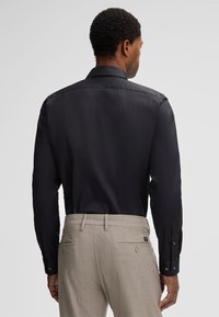 Uomo con capelli ricci corti che indossa una maglietta nera a maniche lunghe e pantaloni beige chiaro, visto da dietro contro uno sfondo semplice.