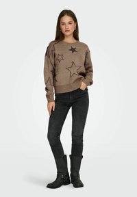 Brązowy sweter dzianinowy z czarnymi gwiazdkami, z długimi rękawami i ściągaczem na dole, w połączeniu z ciemnymi rurkami dżinsowymi i czarnymi wysokimi kozakami.