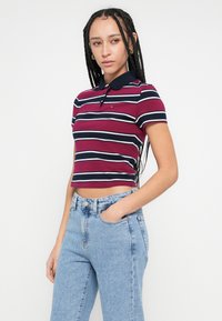 Kratké polo tričko s krátkym rukávom v bordovej farbe s vodorovnými bielymi a námorníckymi pruhmi, s námorníckym golierom a jemným logom. Kombinované so svetlomodrými džínsami.