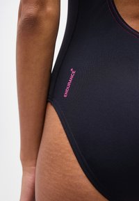 Κοντινό πλάνο μαύρου μαγιό με ροζ ραφές και τη λέξη "ENDURANCE+" τυπωμένη κοντά στο ισχίο πάνω σε σκούρο δέρμα ατόμου.