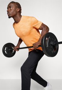 Orange, struktureret t-shirt med et subtilt mønster, kombineret med sorte træningsbukser og hvide sneakers, mens der løftes en vægtstang med vægte.