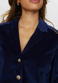 Mornarsko modra kordura blazerka z izrezanim ovratnikom, ki vključuje dve veliki gumb in subtilno teksturo. Povečan posnetek ovratnika in zgornjih podrobnosti tkanine.