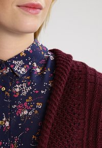 Burgunderfarbener Strickcardigan mit offener Webstruktur, über einer navyblauen Bluse mit Kragen, die ein florales Muster in Pink, Gelb und Weiß zeigt.