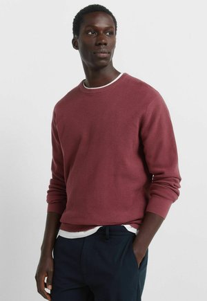 Maglione bordeaux a coste con maniche lunghe e scollo tondo, indossato sopra una t-shirt bianca. Tessuto strutturato, vestibilità slim, abbinato a pantaloni scuri.