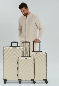 Un homme en tenue beige se tient derrière et tient les poignées de trois valises métalliques beiges avec un motif en chevrons sur un fond gris.