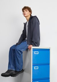 Grauer Reißverschluss-Hoodie über einem grauen T-Shirt, blaue Jeans und schwarze Sneakers; sitzt auf einem grauen Aktenschrank mit blauen Schubladen.