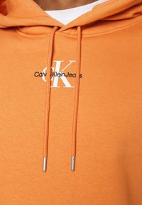 Felpa arancione realizzata in tessuto morbido con cordino e punte in argento. La parte frontale presenta un logo bianco con la scritta "Calvin Klein Jeans".