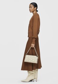 Cardigan de punto marrón y falda midi combinados con botas altas beige. El bolso es de color crema con un patrón texturizado beige y una correa de cadena plateada.