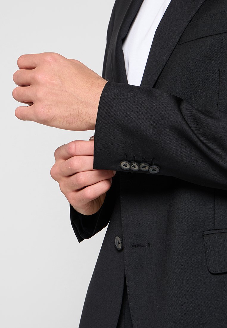 Man die de manchet van een zwart colbert met vier knopen aanpast, draagt een wit shirt eronder, tegen een effen lichte achtergrond.