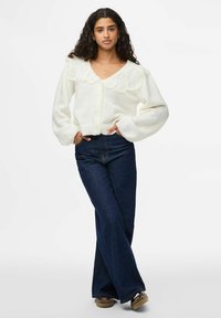 Vit stickad kavaj med V-hals och vågiga detaljer runt axlarna, i kombination med mörkblå utsvängda jeans och bruna skor.