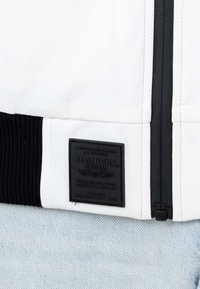 Giubbotto bomber bianco con orlo a coste nero e un'etichetta quadrata nera con testo in rilievo, posizionata vicino alla chiusura con zip.