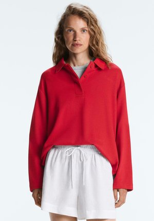 Kvinde med langt bølget hår iført en løs rød langærmet skjorte med krave over en grå top og hvide shorts med snøre, vendt mod fronten.