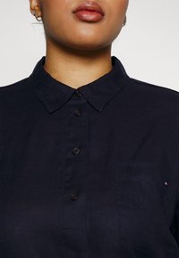 Chemise en lin bleu marine avec un devant à boutons, un col et une poche poitrine simple. Elle présente des boutons noirs et un petit détail de logo sur la poche.