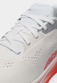 Λευκό αθλητικό παπούτσι με λευκά κορδόνια, διάτρητο επάνω μέρος, γκρι οπές και κόκκινο λογότυπο "ACT" στο πλάι κοντά σε κόκκινη σόλα με απόχρωση.