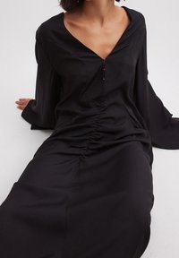 Černé šaty s dlouhými rukávy, V-výstřihem a na přední straně nařasením. Vyrobeno z hladké tkaniny s volným střihem a jemným leskem.