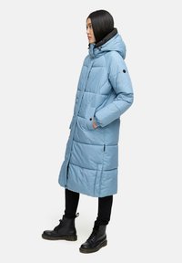 Svijetloplavi pufasti kaput s kapuljačom, s prepoznatljivim prošivenim dizajnom, rebrastim manžetama i bočnim džepovima. Uparen s crnim hlačama i čizmama.