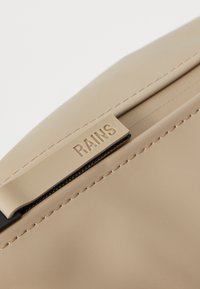 Beige silikontaske nærbilde, med en svart glidelås og preget logo. Glatt tekstur med synlige sømdetaljer langs kanten.