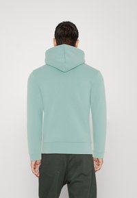 Felpa con cappuccio verde menta dal tessuto liscio e maniche a costine, abbinata a pantaloni verde scuro, visibile da un retro casual.