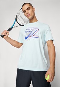 Camiseta de algodón azul claro con un diseño gráfico de Nike en púrpura y rojo. El modelo sostiene una raqueta de tenis y una pelota de tenis verde.
