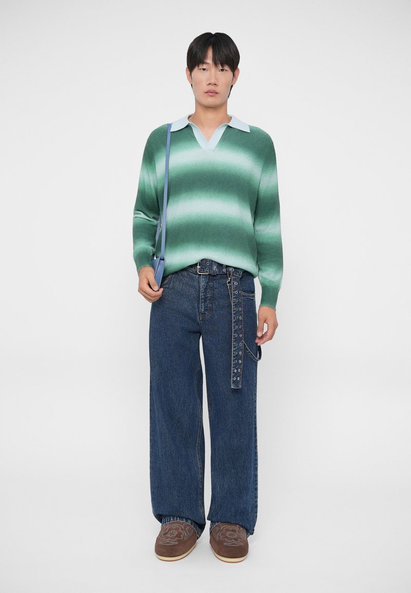 Maglione a maglia con gradiente verde e colletto blu chiaro, abbinato a jeans in denim blu scuro a gamba larga. Gli accessori includono una borsa blu e una cintura con hardware.