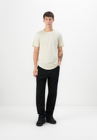 Camiseta de manga corta color beige claro con dobladillo redondeado, combinada con pantalones de pierna ancha negros y zapatillas negras, de pie frente a un fondo blanco.