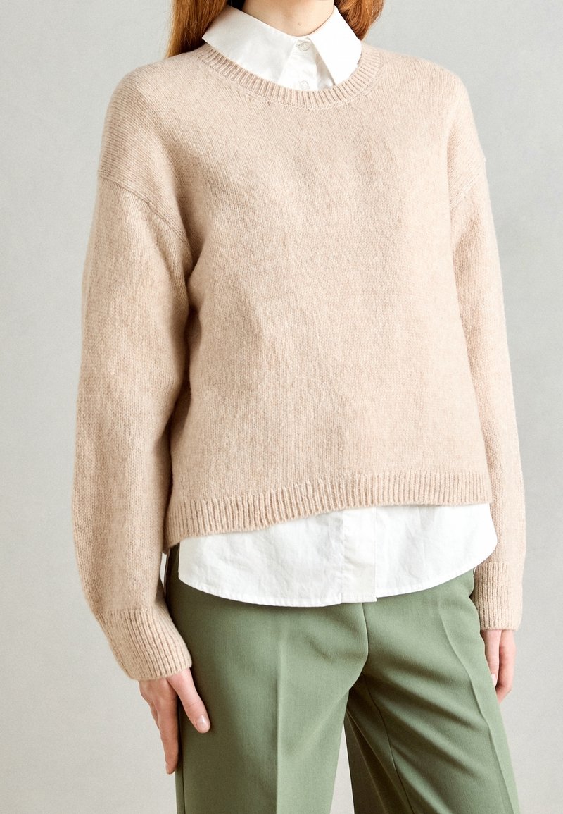 Femme portant un pull en maille beige sur une chemise blanche boutonnée, associée à un pantalon vert clair ajusté, debout devant un fond neutre.