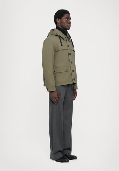Olivgrüne Kapuzenjacke mit zwei vorderen Taschen, Knopfverschluss und Kordelzügen. Getragen über dunklen grauen Hosen und schwarzen Schuhen.