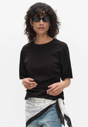 Vrouw met krullend haar, zwarte zonnebril, een zwart shirt met korte mouwen, blauwe jeans en een wit sjaaltje met witte stippen dat in de taille is gestrikt.